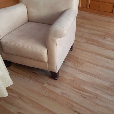 Beiger Sessel auf Boden in Holzoptik. Seite eines Tisches mit weißem Tuch sichtbar.