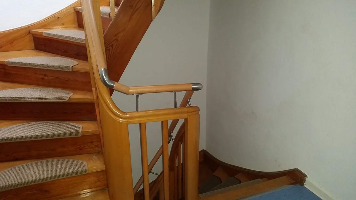 Geschwungene Holztreppe mit Teppichstufen und Holzhandlauf vor einer weißen Wand.