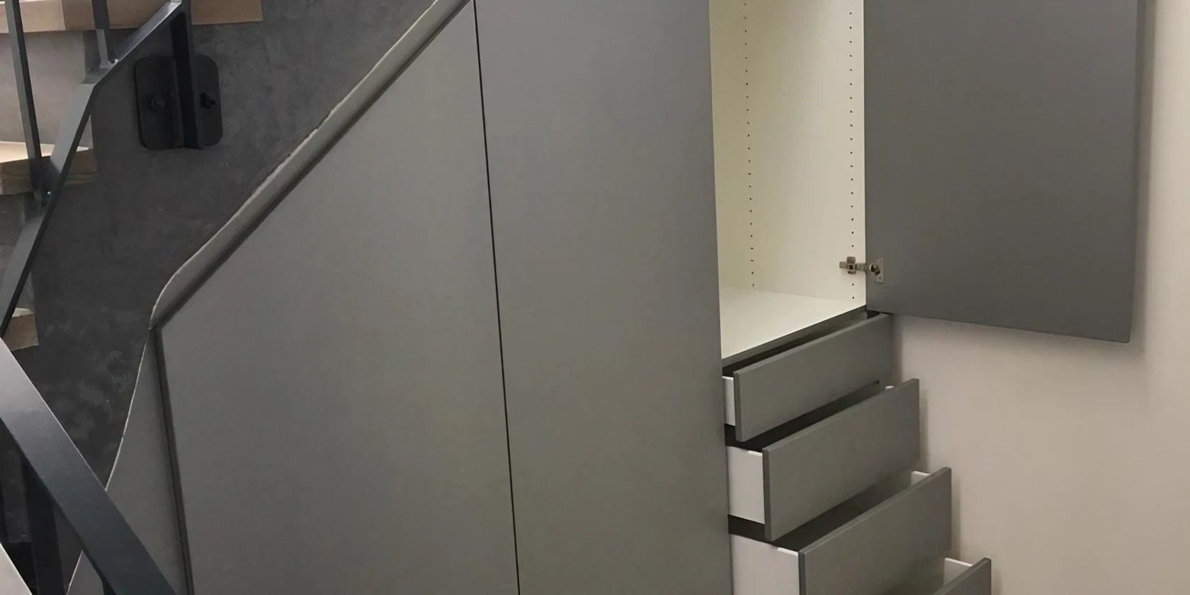 Ein graues, in die Wand unter einer Treppe eingebautes Aufbewahrungsmöbel mit drei Schubladen und einer offenen Tür.
