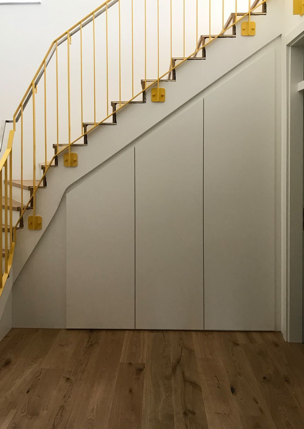 Hellgrauer Einbauschrank unter einer Treppe mit gelbem Geländer, in einem Raum mit Holzboden.
