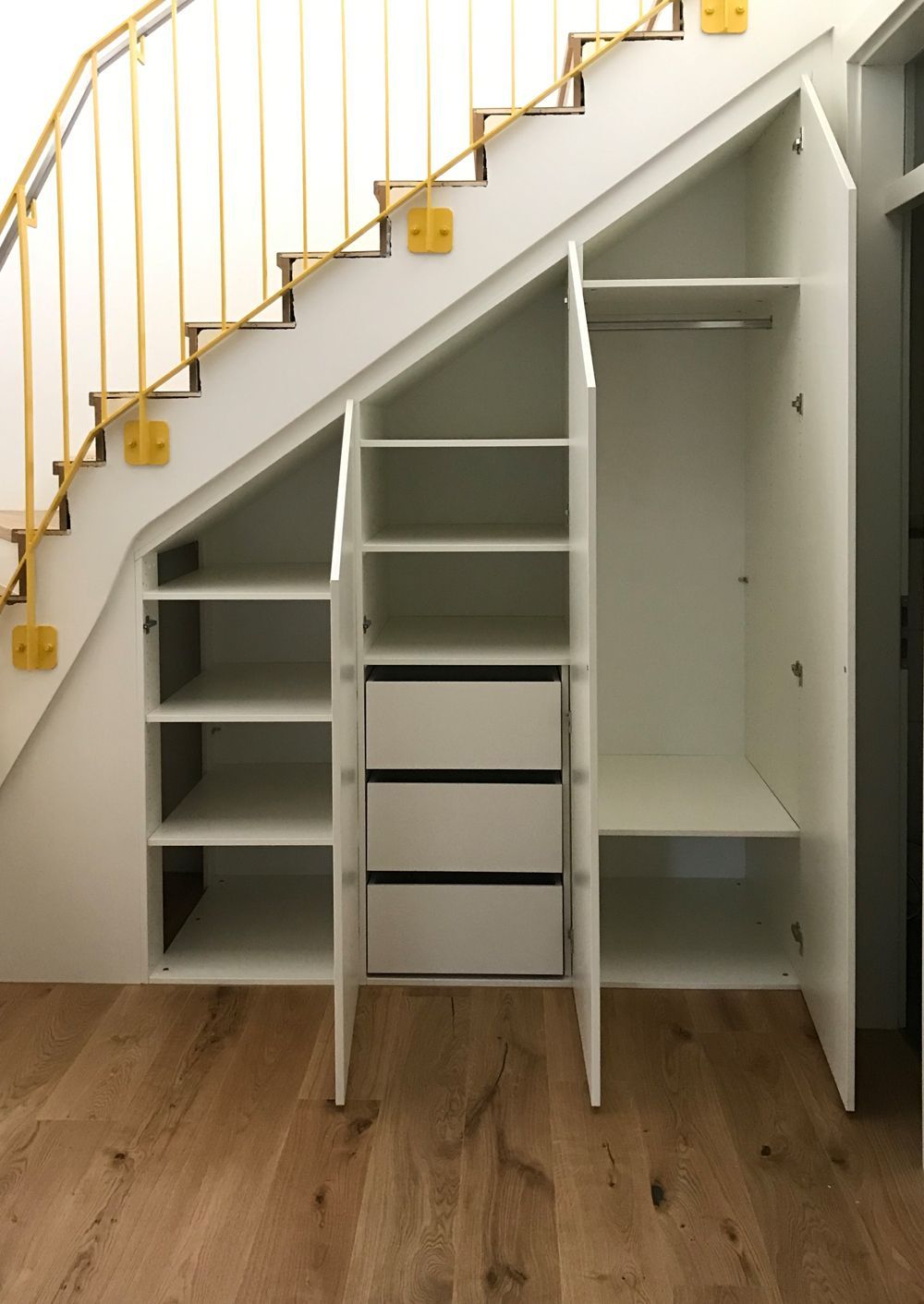 Weißer Einbauschrank unter der Treppe mit Regalen, Schubladen und Kleiderschrank. Holzboden, gelbes Treppengeländer.