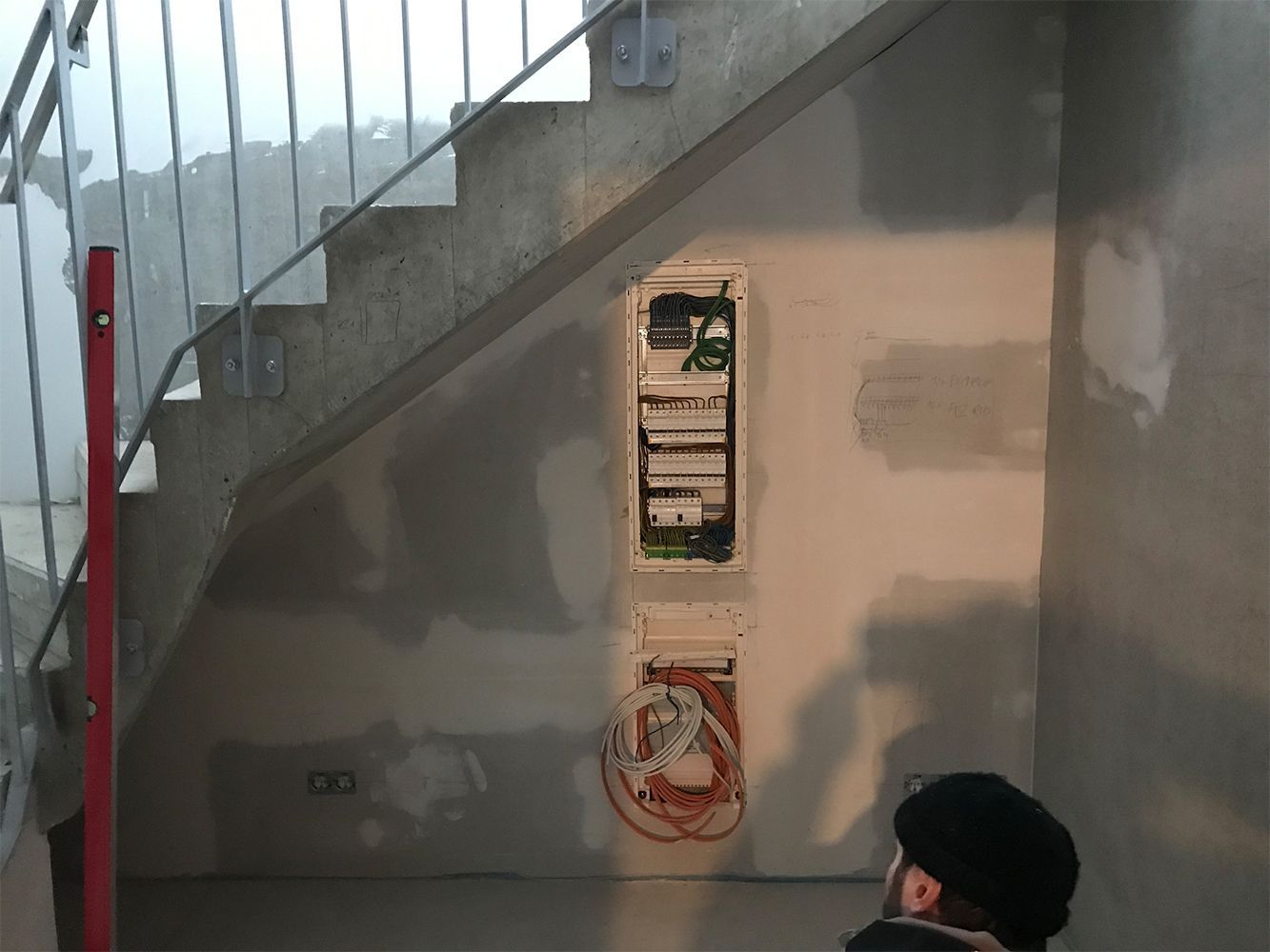 Elektrischer Schaltschrank und Verkabelung unter einer Treppe. Eine Person mit Hut betrachtet ihn.