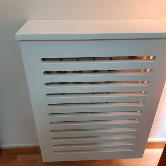 Una cubierta de radiador blanca está colocada sobre un piso de madera al lado de una pared.