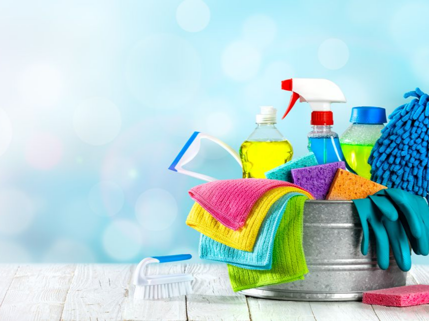 Produits de nettoyage dans un seau en métal : flacons pulvérisateurs, éponges, chiffons, brosse, gants, sur fond bleu clair.