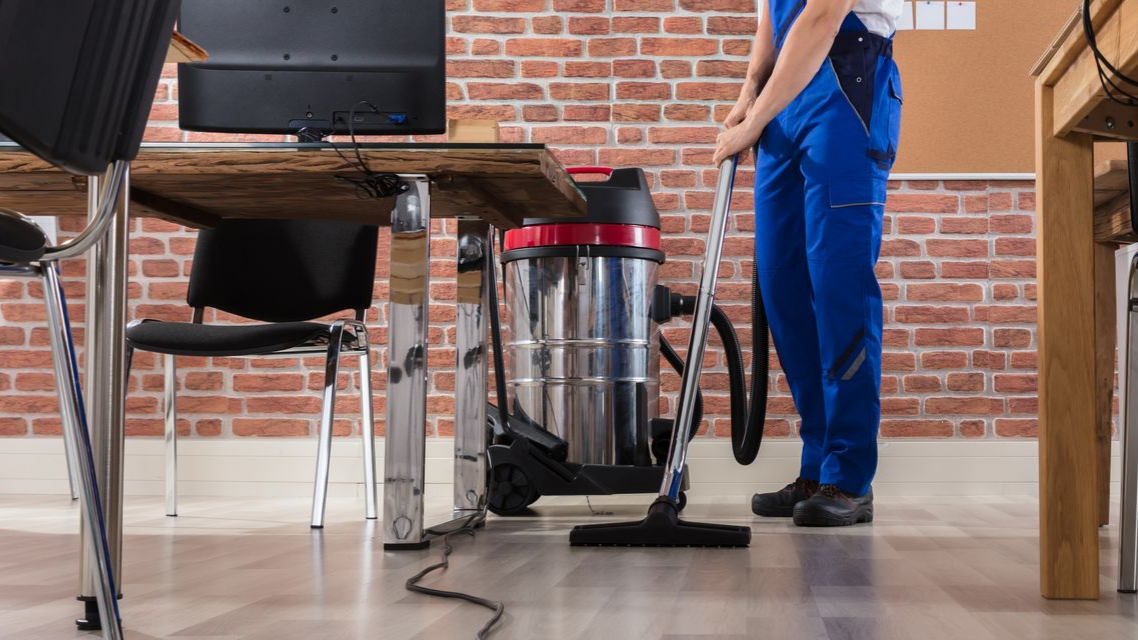 Une personne en salopette bleue passe l'aspirateur sur le sol d'un bureau avec un aspirateur eau et poussière.