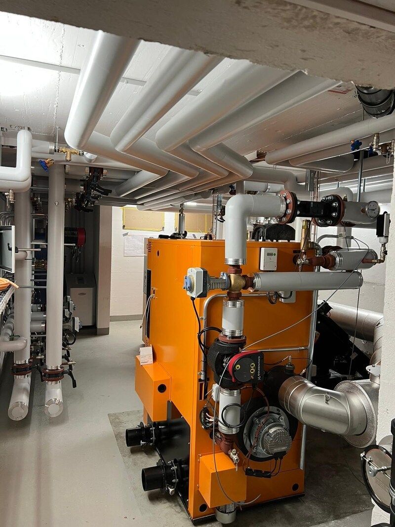 Eine große orangefarbene Maschine steht in einem von Rohren umgebenen Raum.