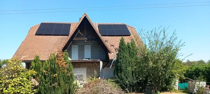 Maison au toit pentu et aux panneaux solaires, entourée d'arbres et de buissons sous un ciel bleu limpide.