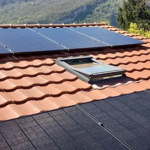 Des panneaux solaires et un puits de lumière sont installés sur un toit de tuiles rouges, avec des montagnes en arrière-plan.