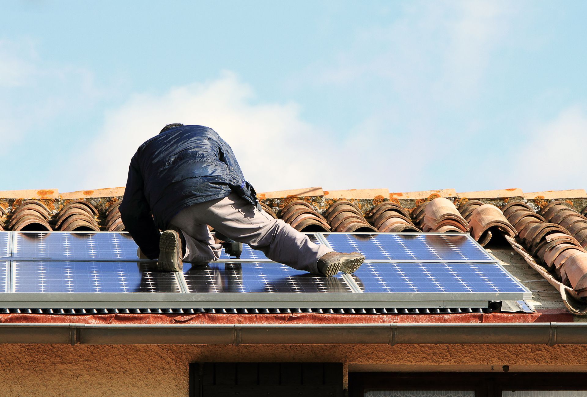 Une personne installe des panneaux solaires sur un toit de tuiles sous un ciel bleu.