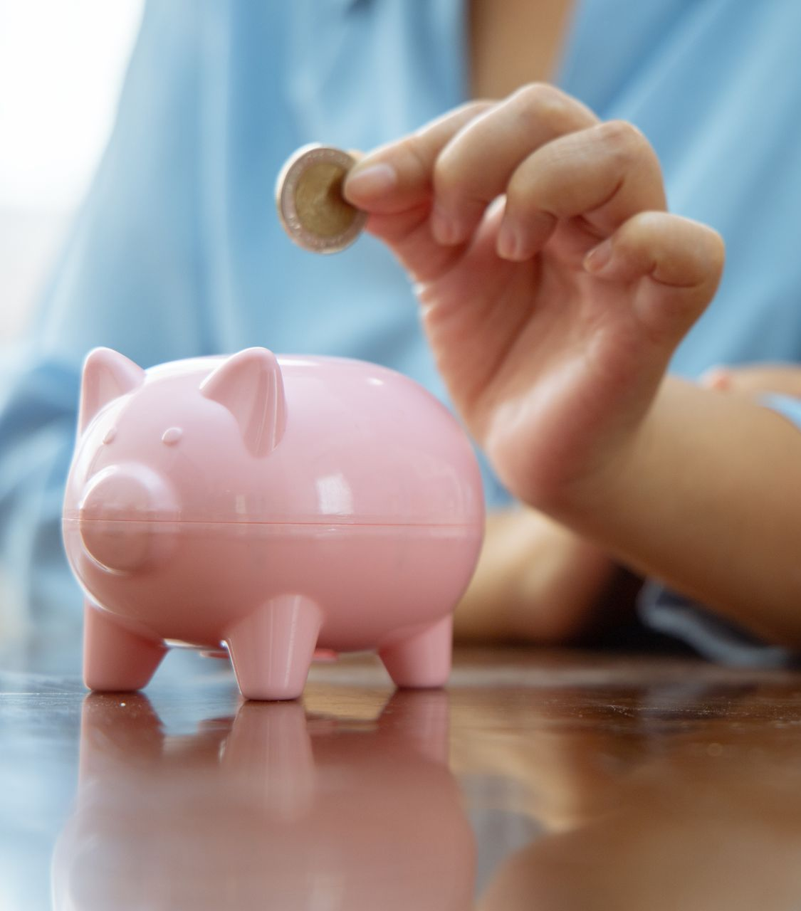 Une personne place une pièce dans une tirelire rose posée sur une table en bois.