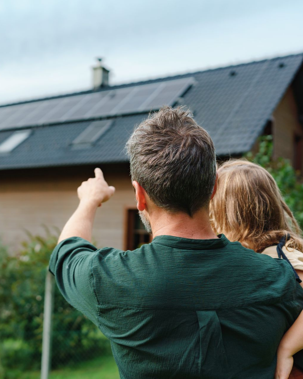 Un homme montre du doigt des panneaux solaires sur le toit d'une maison tout en tenant un enfant dans ses bras.
