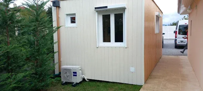 Petit bâtiment préfabriqué beige en bordure de trottoir, avec une fenêtre, un climatiseur et un véhicule stationné à proximité.