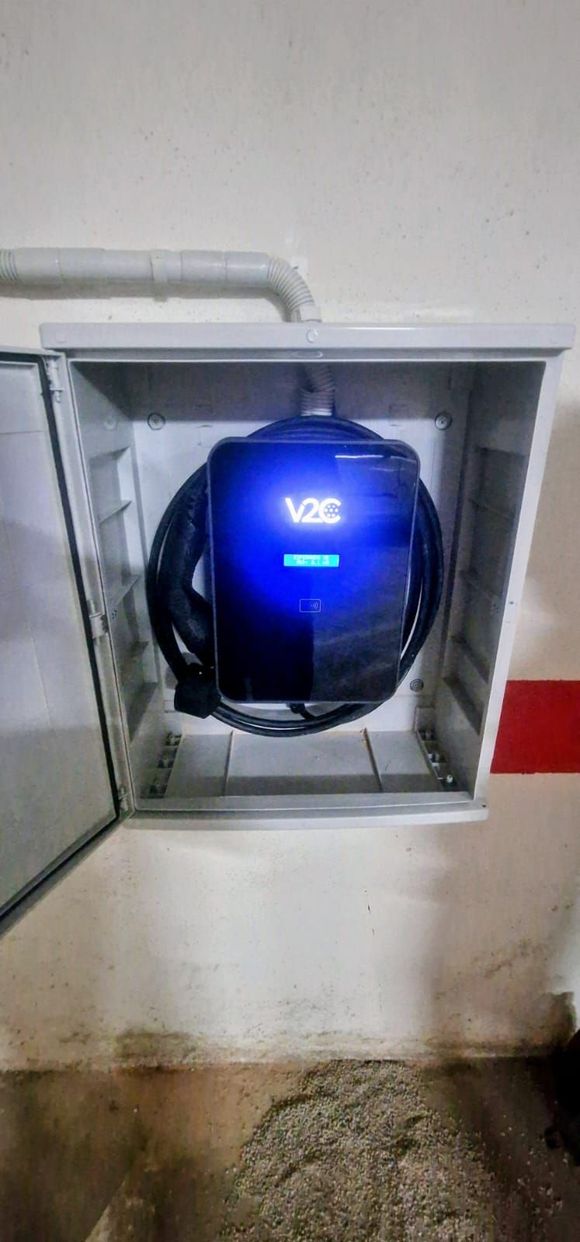 Una estación de carga para vehículos eléctricos montada en la pared. Está encerrada en una caja metálica gris y tiene una pantalla azul que muestra