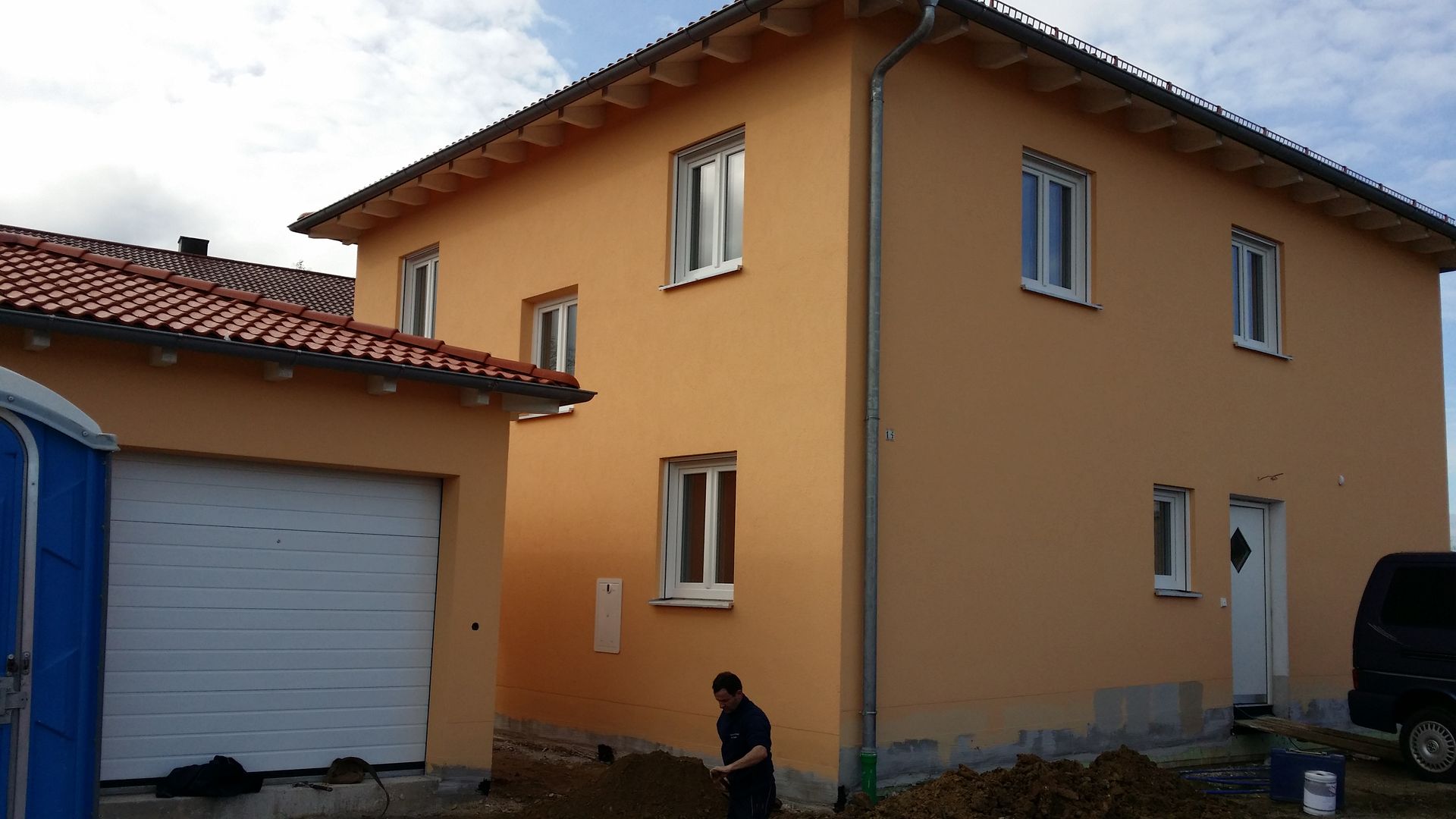 Zweistöckiges orangefarbenes Haus mit Garage und eine Person, die draußen an der Mauer kauert.