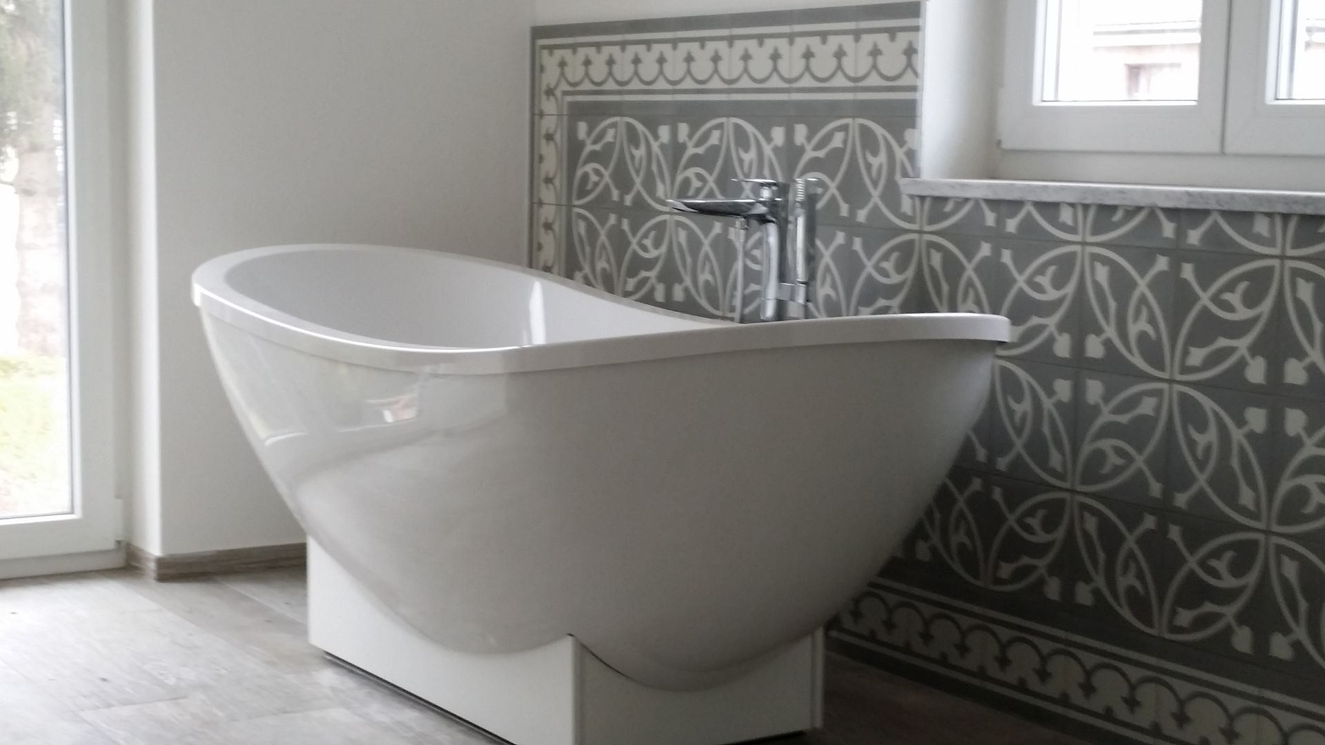 Weiße, freistehende Badewanne in einem hellen Badezimmer mit gemusterter Fliesenwand und Fenster über dem Waschbecken