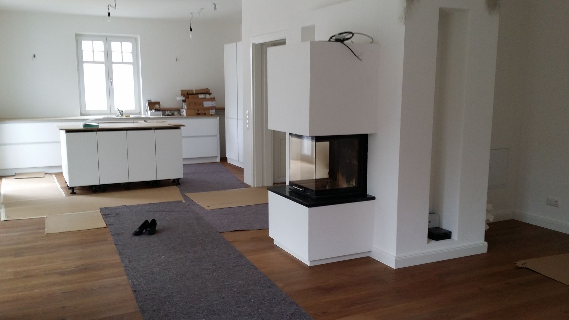 Helles, leeres Wohnzimmer mit weißem Kamin, Holzböden und Kücheninsel im Hintergrund