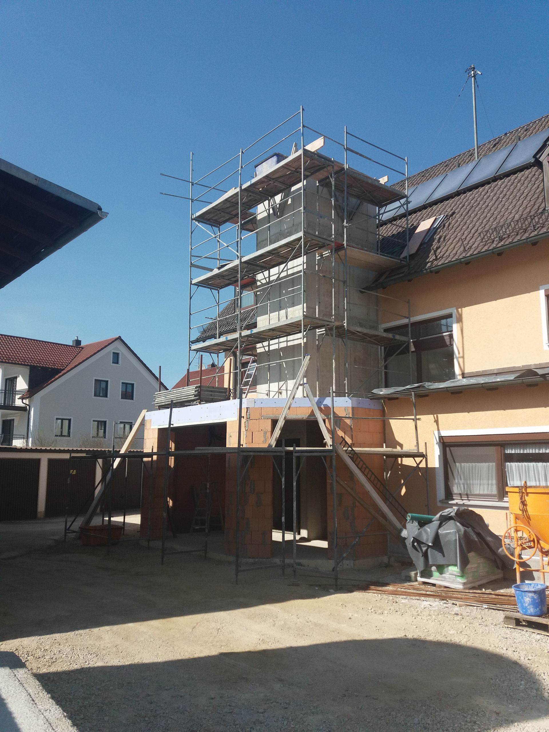 Hausfassade mit Gerüst in einem sonnigen Innenhof, mit Zaun und nahegelegenen Nachbarhäusern.