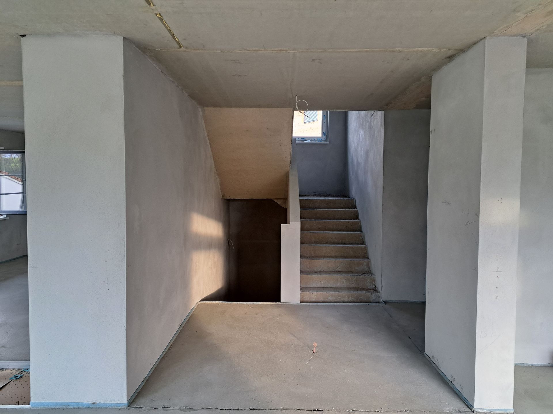 Leerer Eingangsbereich aus Beton mit zentralem Treppenhaus und weißen Säulen, der von einem kleinen Fenster darüber beleuchtet wird.