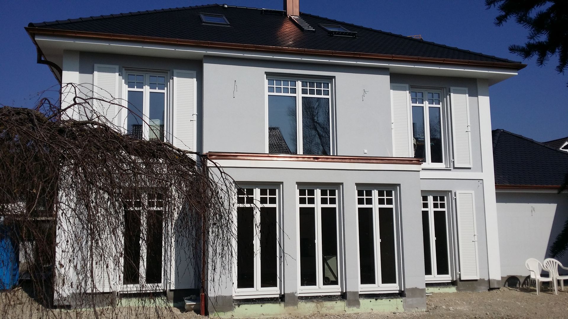 Zweistöckiges weißes Haus mit dunklem Dach, großen Fenstern und Flügeltüren in einem sonnigen Garten