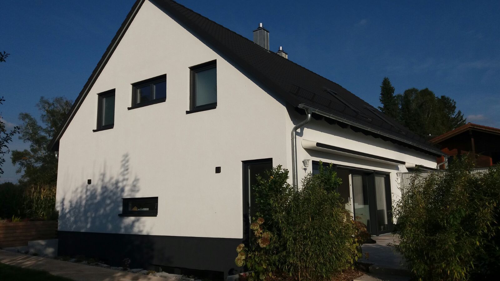 Weißes zweistöckiges Haus mit schwarzem Dach und schwarzen Fenstern, von vorne im hellen Sonnenlicht gesehen.