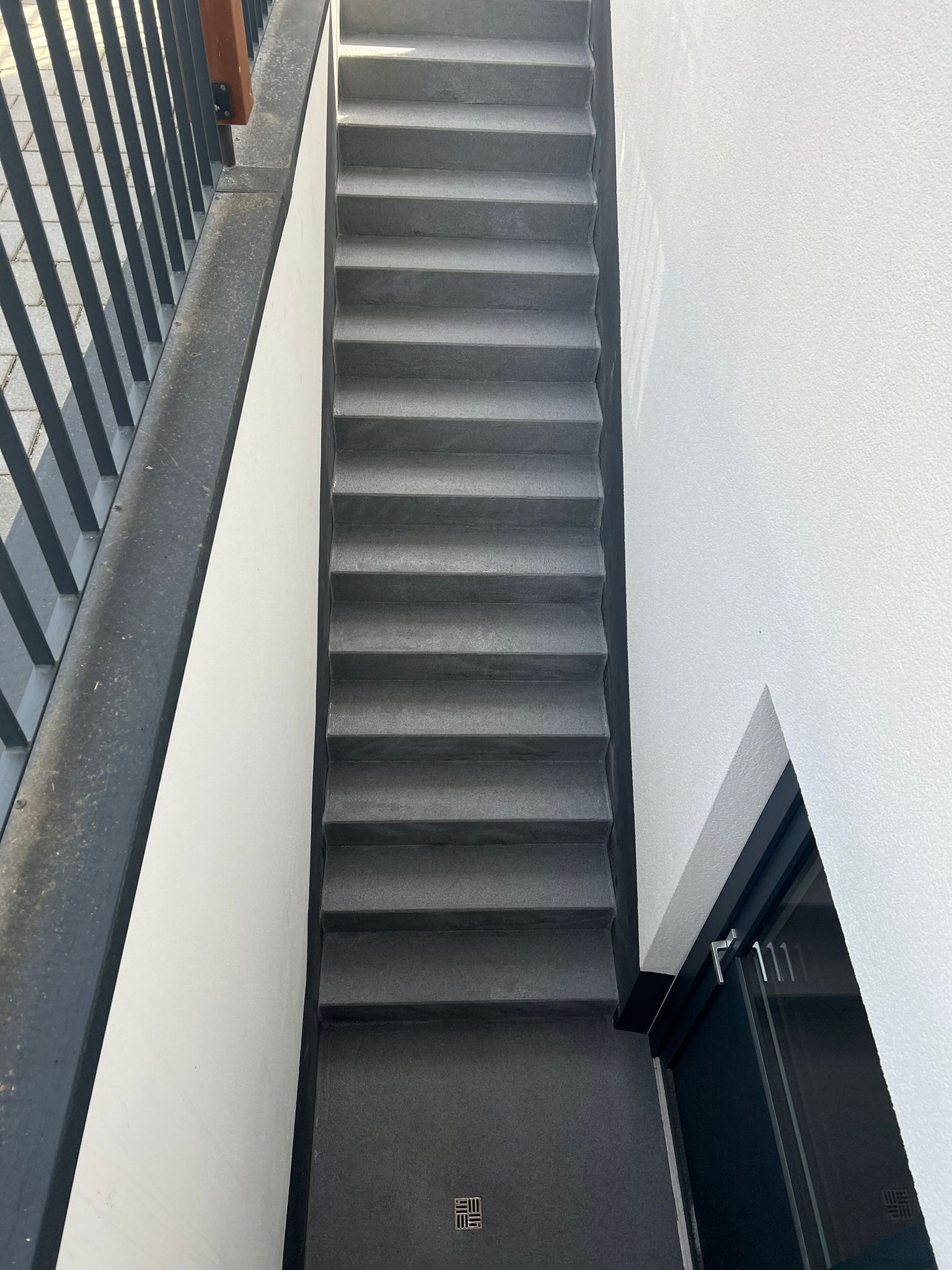 Schmale, graue Treppe mit Metallgeländer und weißen Wänden, vom unteren Treppenabsatz aus gesehen.
