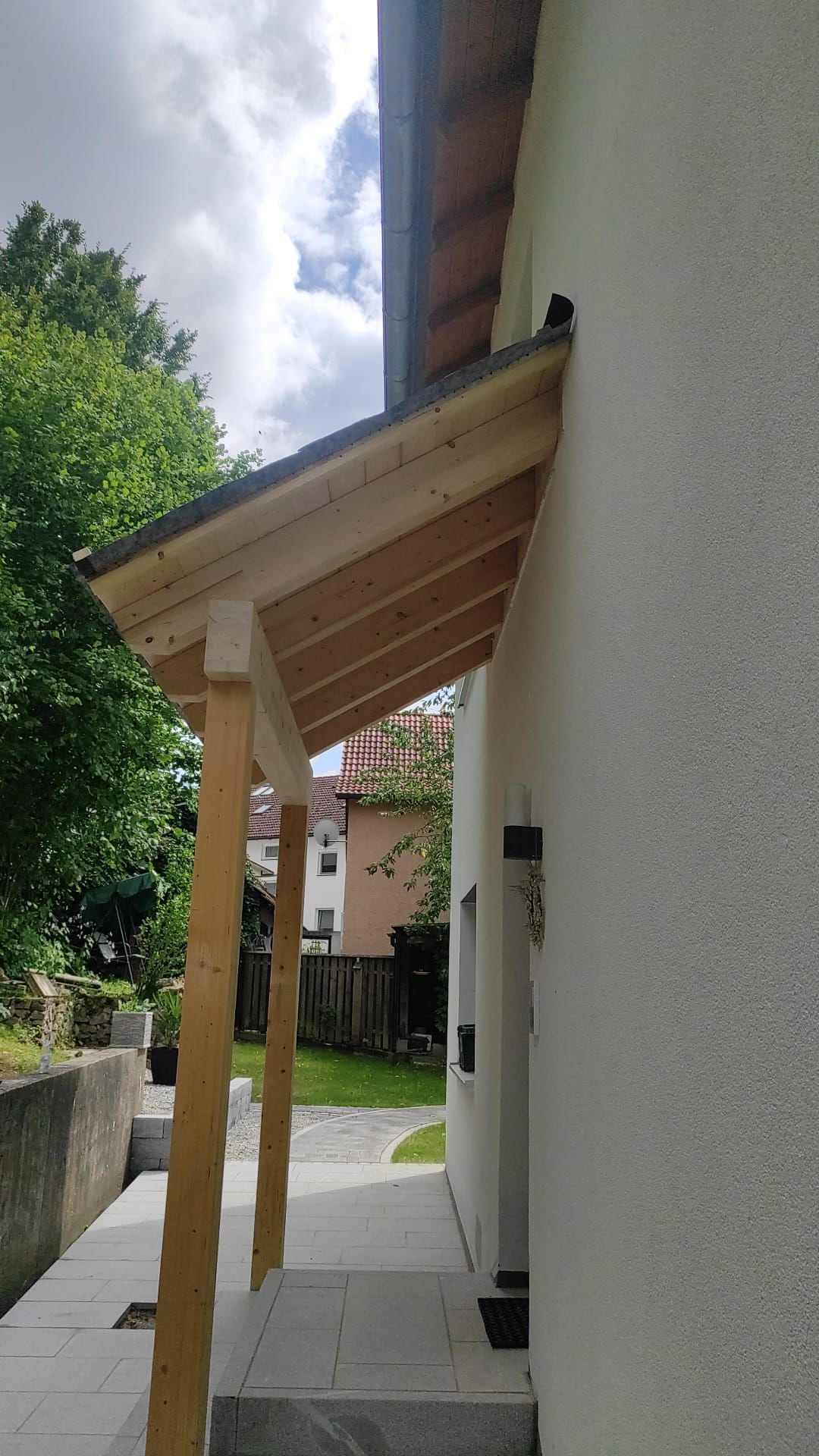 Seitenansicht eines weißen Hauses mit hölzernem Vordach und Pfosten entlang eines gepflasterten Gehwegs.