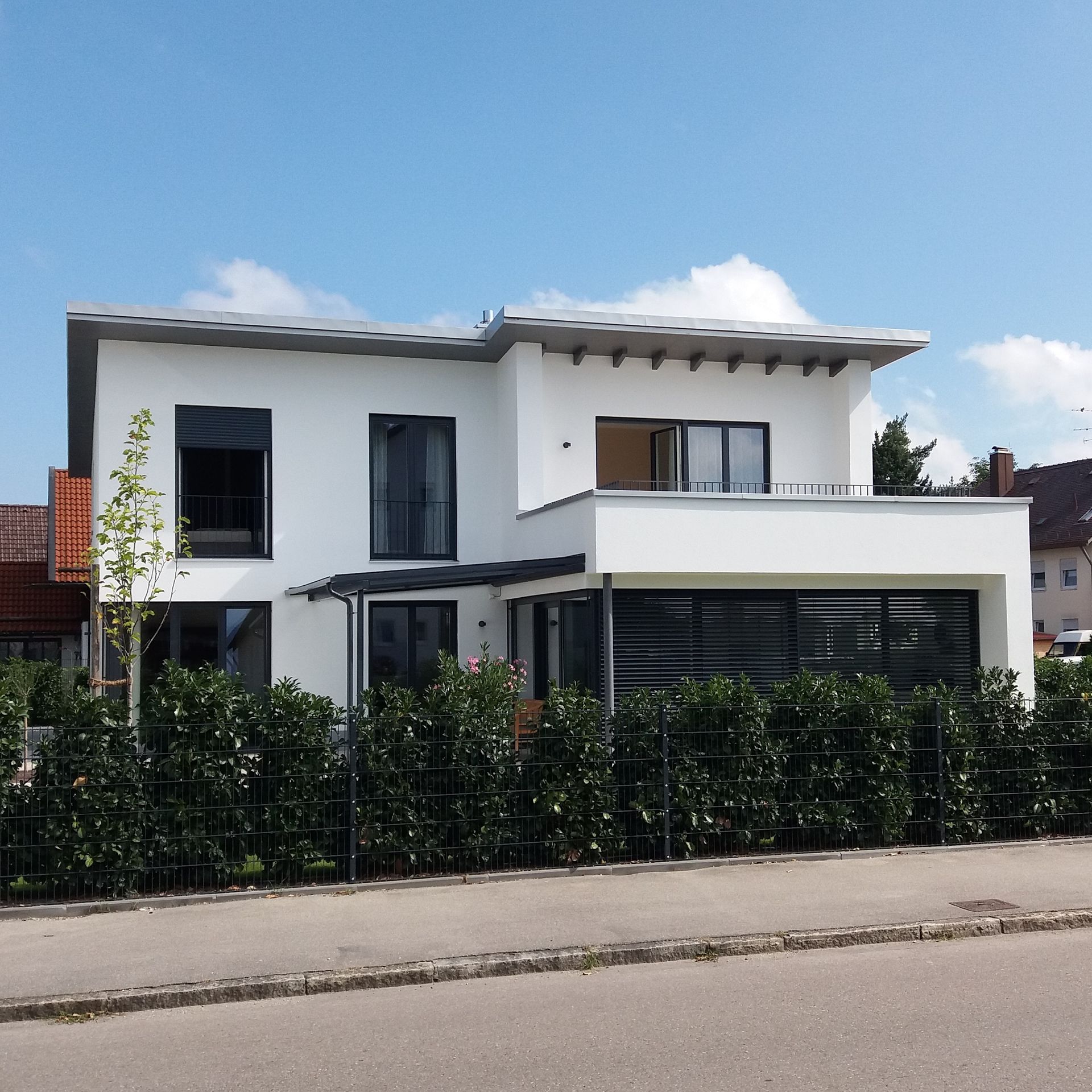 Modernes, weißes zweistöckiges Haus mit großen Fenstern und einer Hecke im Vorgarten an einer ruhigen Straße