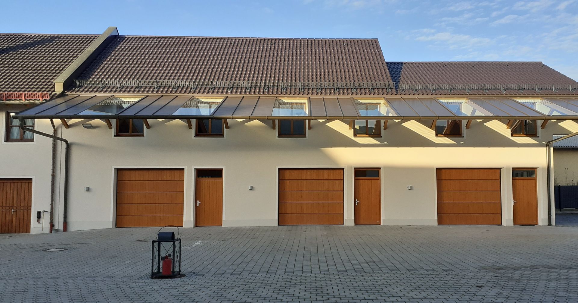 Eine Reihe weißer Garagentore unter einem Ziegeldach in einem sonnigen Innenhof, davor ein kleiner Mülleimer.