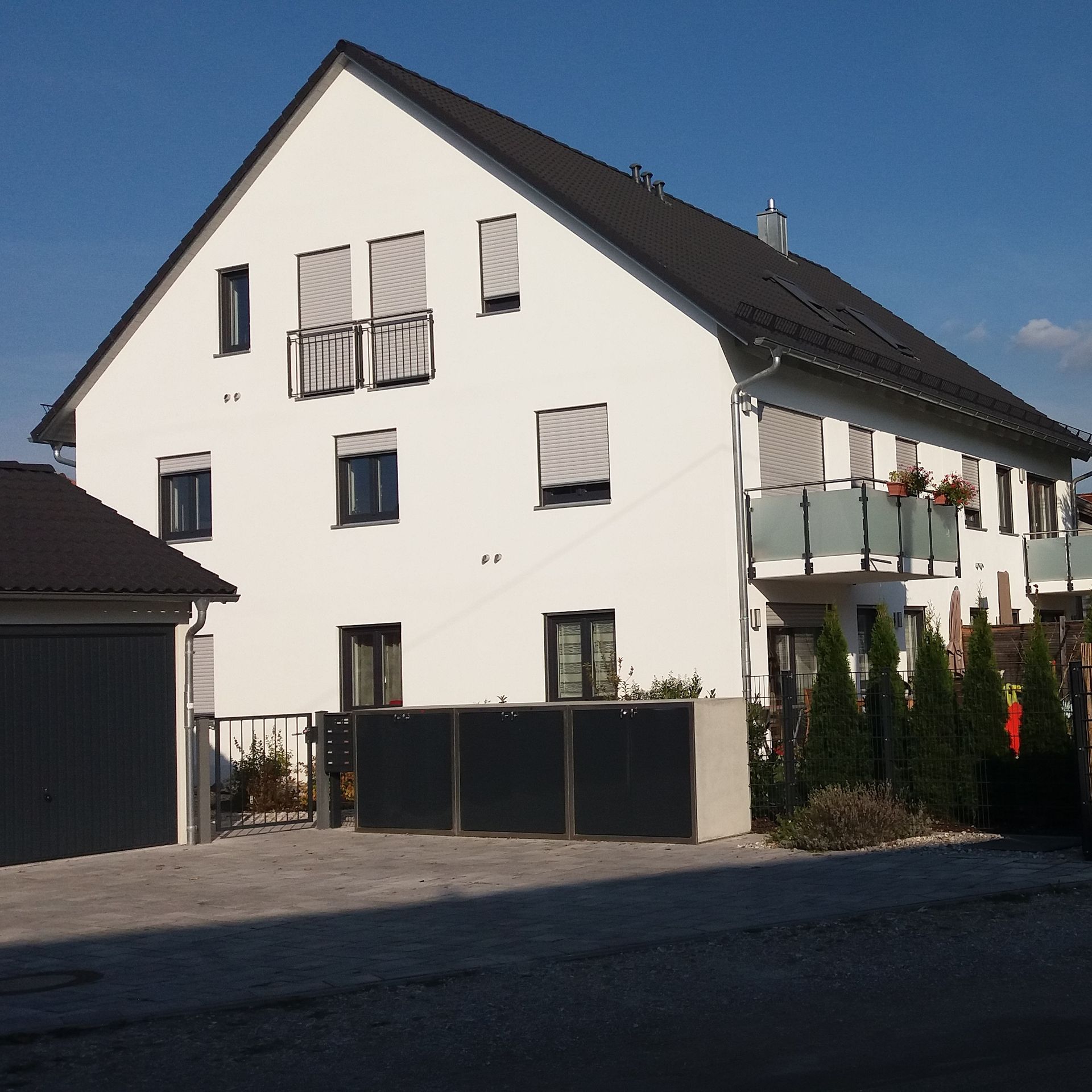 Weißes zweistöckiges Haus mit Balkon, dunklem Dach und Garage in einer sonnigen Wohnstraße