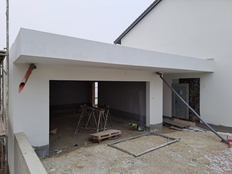 Eine im Bau befindliche weiße Garagenkonstruktion mit großem, offenem Eingang und diagonalem Stützbalken im Freien.