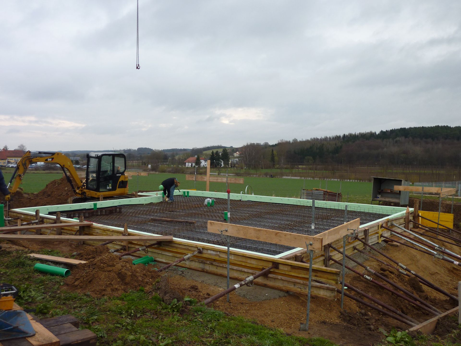 Baustelle mit Bagger, Arbeitern und hölzernen Fundamentschalungen auf schlammigem Boden unter bewölktem Himmel