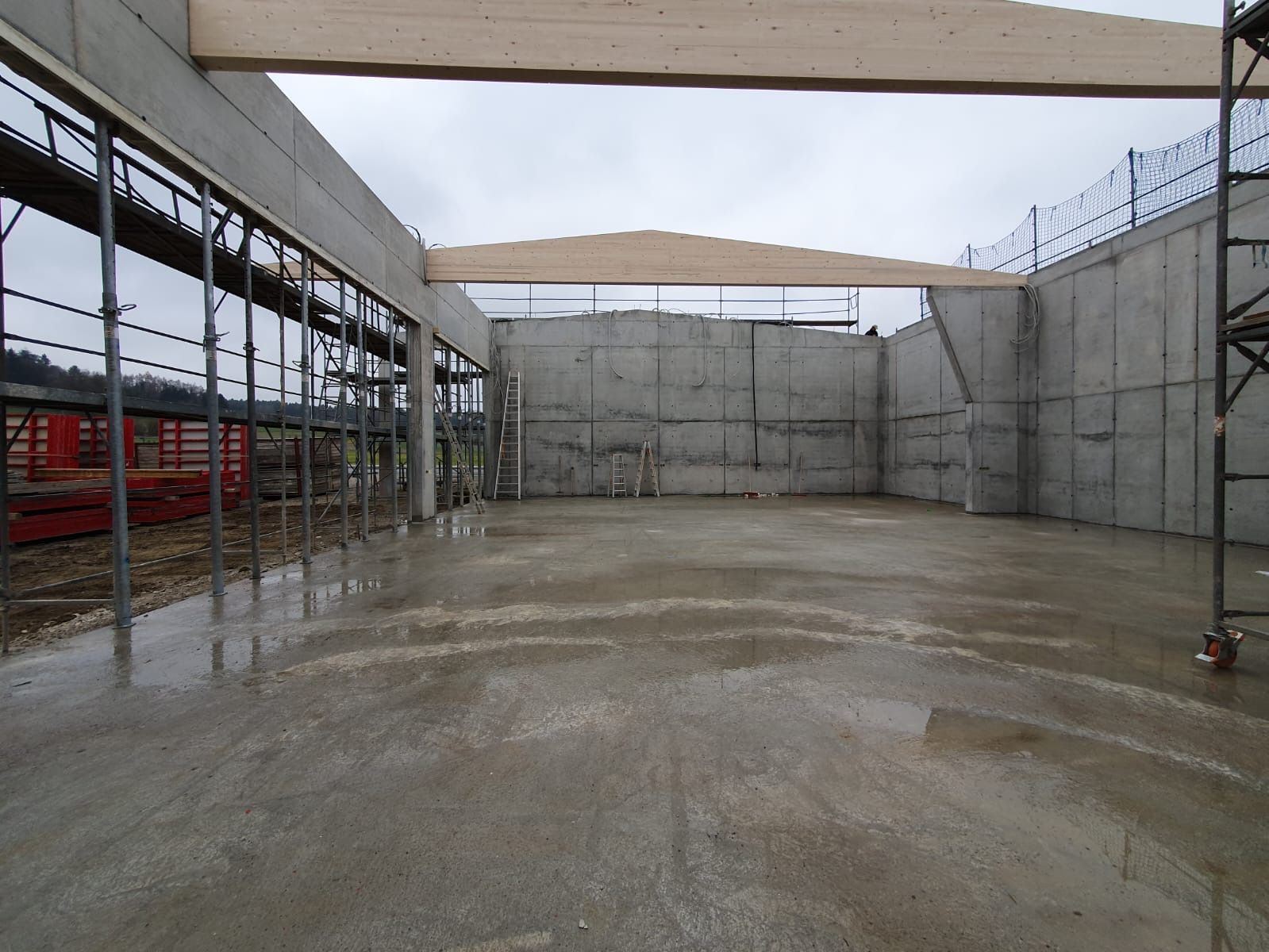 Leere Betonbaustelle mit nassem Boden, grauen Wänden und Stahlkonstruktion unter einem offenen Dach