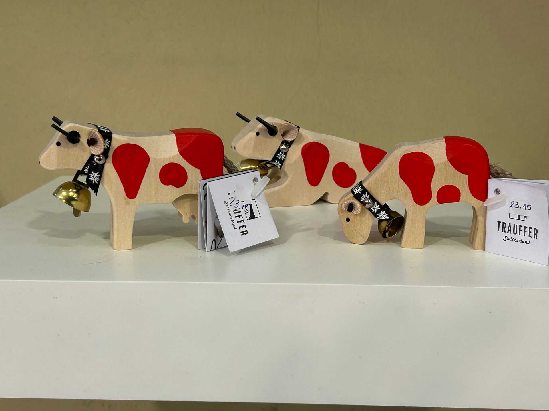Trois figurines de vaches en bois avec des taches rouges, des clochettes et des étiquettes, sur une étagère blanche.