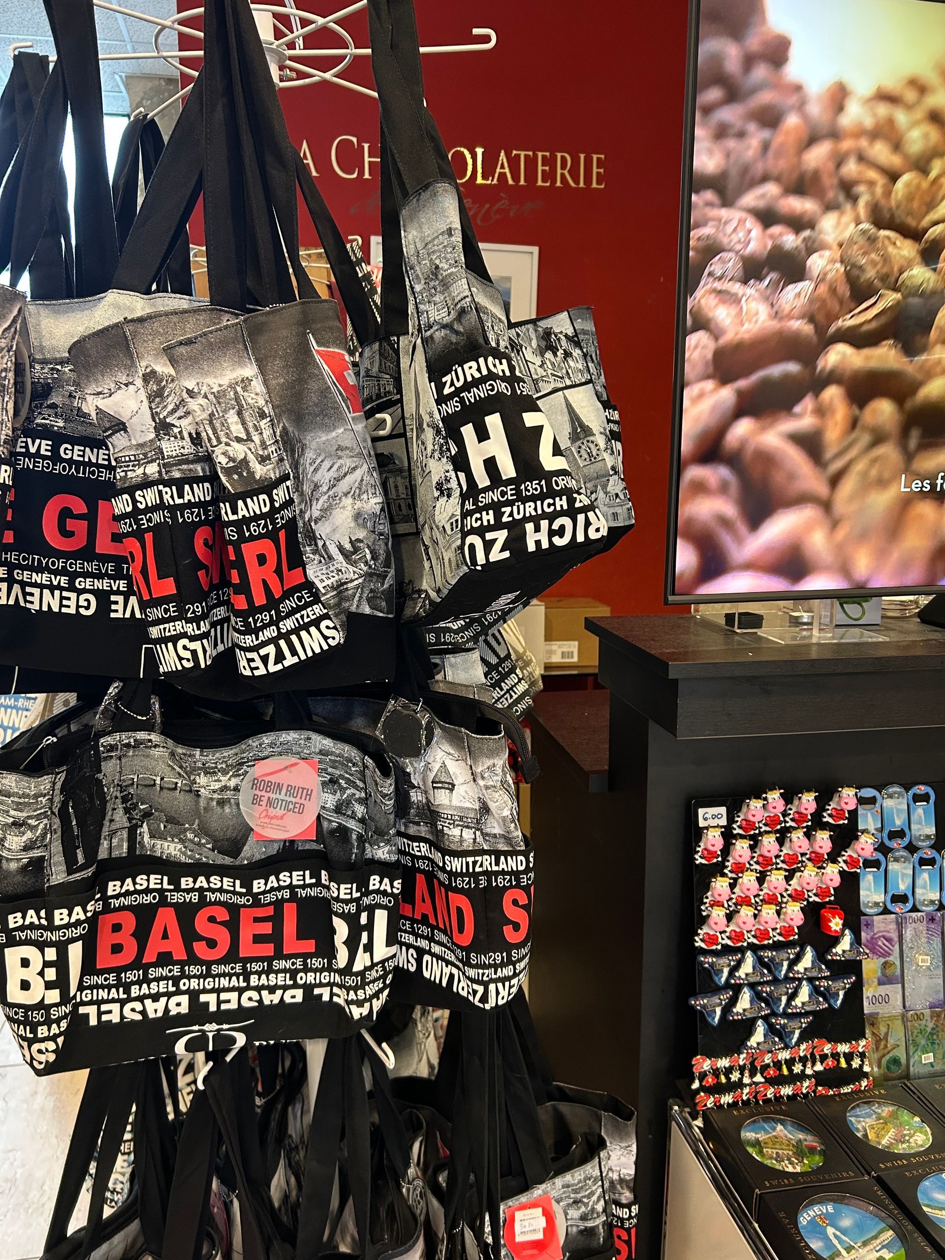 Des sacs fourre-tout avec des noms de villes en rouge et noir, accrochés dans une boutique, peut-être chez un chocolatier.
