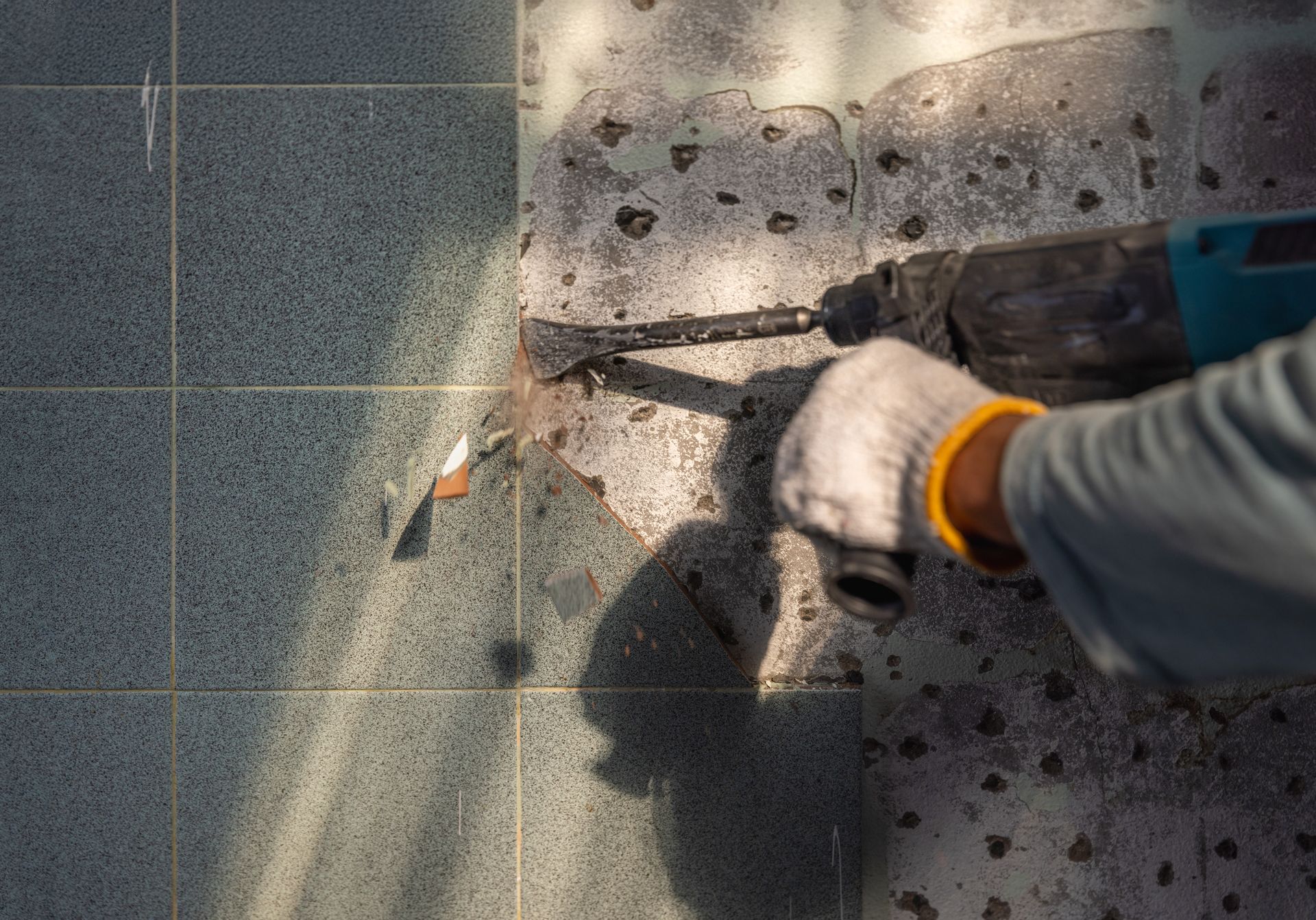Une personne gantée utilise un marteau pour casser du béton à côté d'un sol carrelé et de débris.