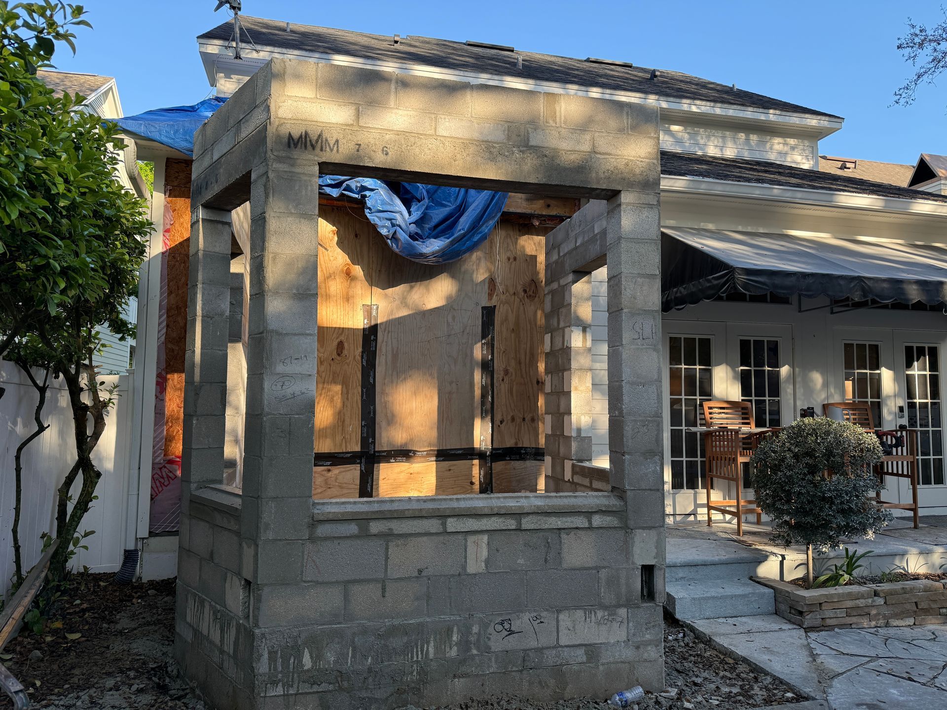 Structure de porche en béton partiellement construite, recouverte d'une bâche bleue, à côté d'une maison dans une cour ensoleillée