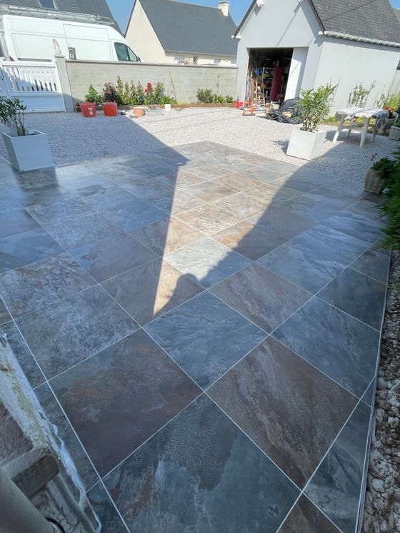 Une terrasse en pierre mène à une maison blanche dont la porte est ouverte et des fleurs en pot baignées de soleil.