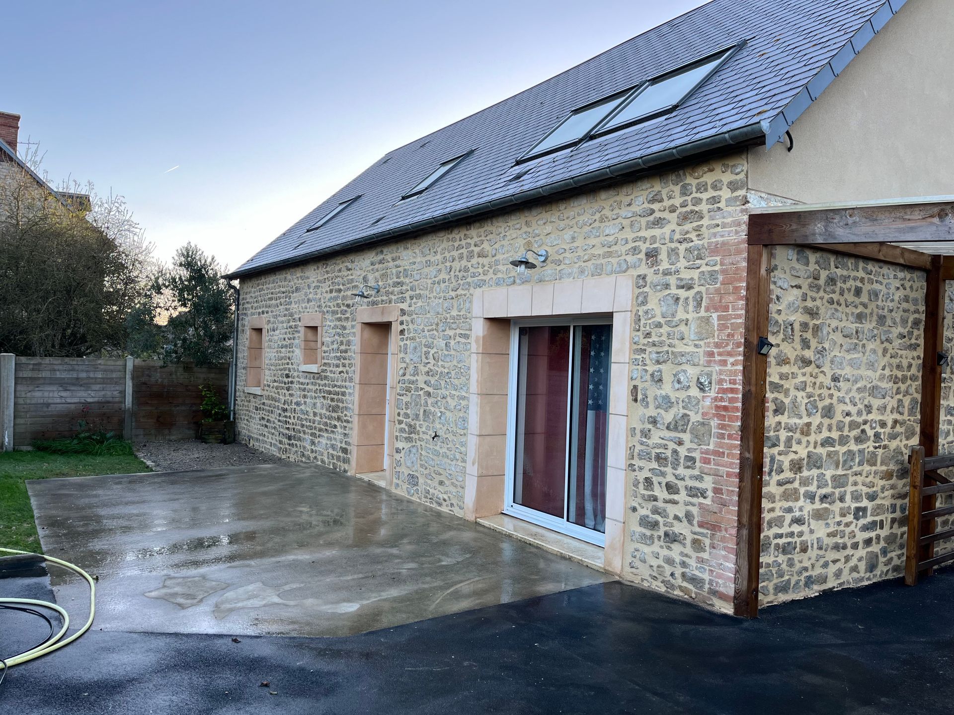 Maison en pierre avec toit en ardoise foncée, portes-fenêtres et allée en béton mouillé donnant sur un jardin gazonné