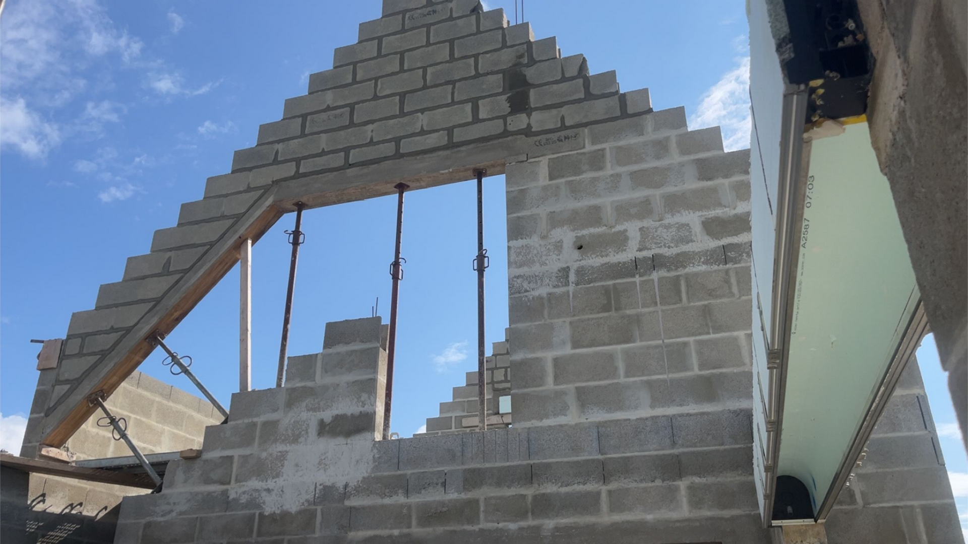 Mur de briques partiellement construit avec une grande ouverture triangulaire et des supports suspendus sous un ciel bleu