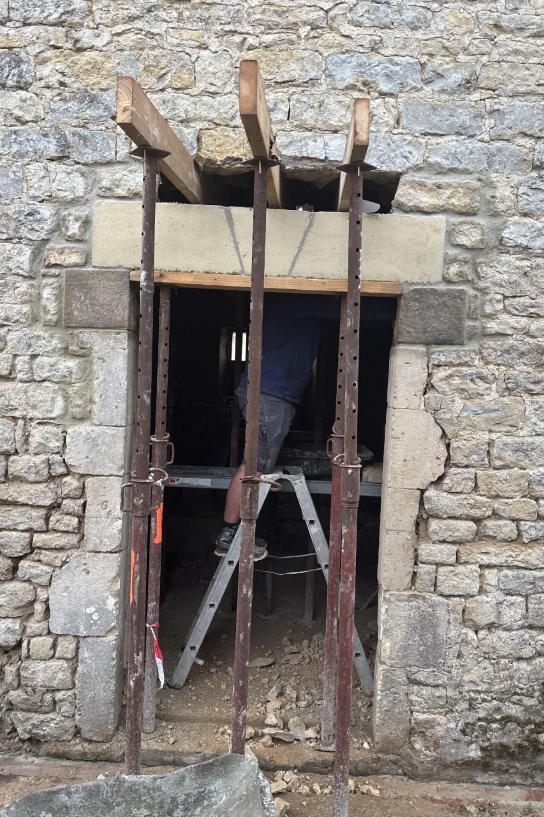 Porte en pierre obstruée par un échafaudage métallique rouillé sur la façade brute d'un bâtiment en pierre.
