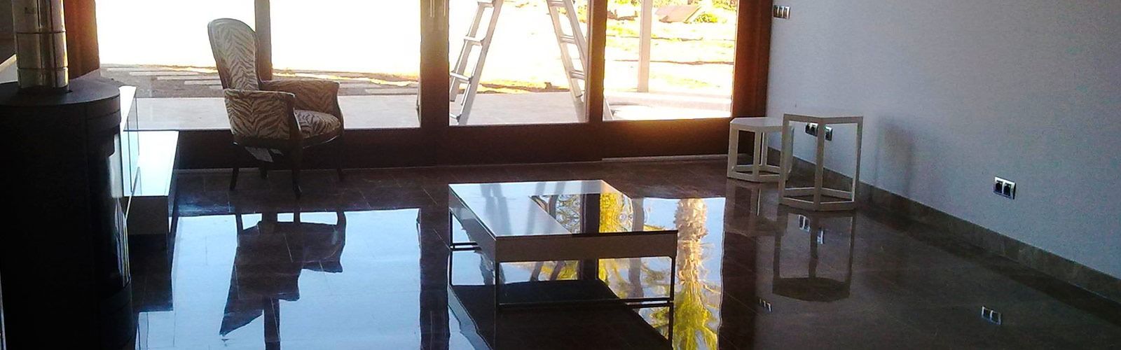 Una habitación con pisos oscuros y brillantes, ventanas grandes y una escalera afuera; muebles visibles.