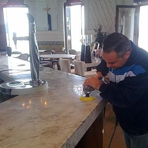 Un hombre pule una barra de mármol con una herramienta eléctrica dentro de un restaurante lleno de luz.