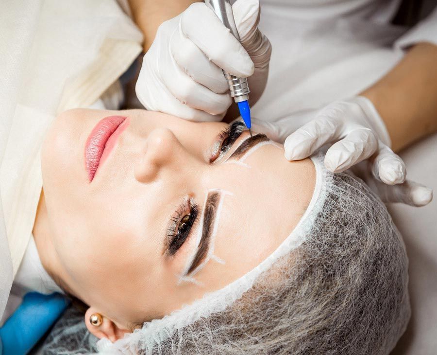 Una técnica, con guantes, utiliza un lápiz de tatuaje cosmético para delinear y rellenar las cejas de una clienta.