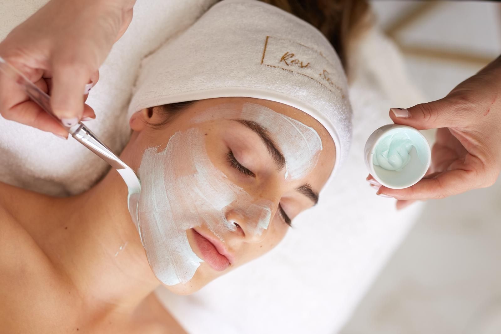 Una persona recibe un tratamiento facial profesional con los ojos cerrados, en el que un técnico le aplica una mascarilla con una brocha.