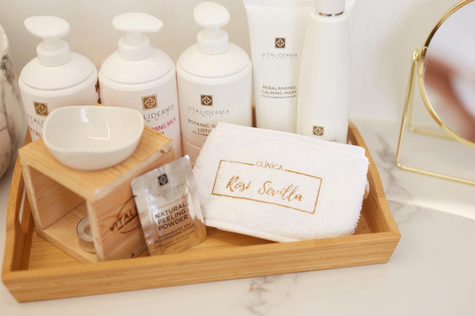 Una bandeja de madera sobre una superficie de mármol contiene productos de baño, un quemador de aceites y una pequeña toalla blanca bordada con