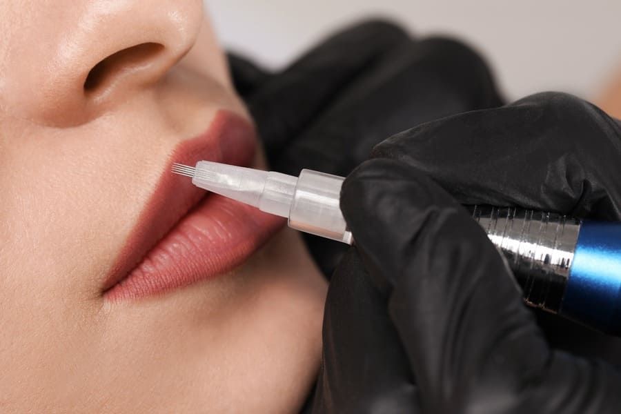 Una persona que lleva guantes negros utiliza un lápiz de tatuaje cosmético para aplicar pigmento en los labios de otra persona.