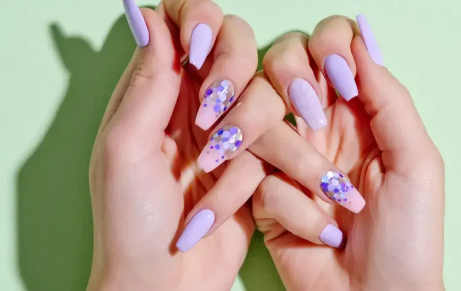 Manos con uñas largas de color lavanda, algunas con degradados de purpurina morada, posando sobre un fondo verde claro.