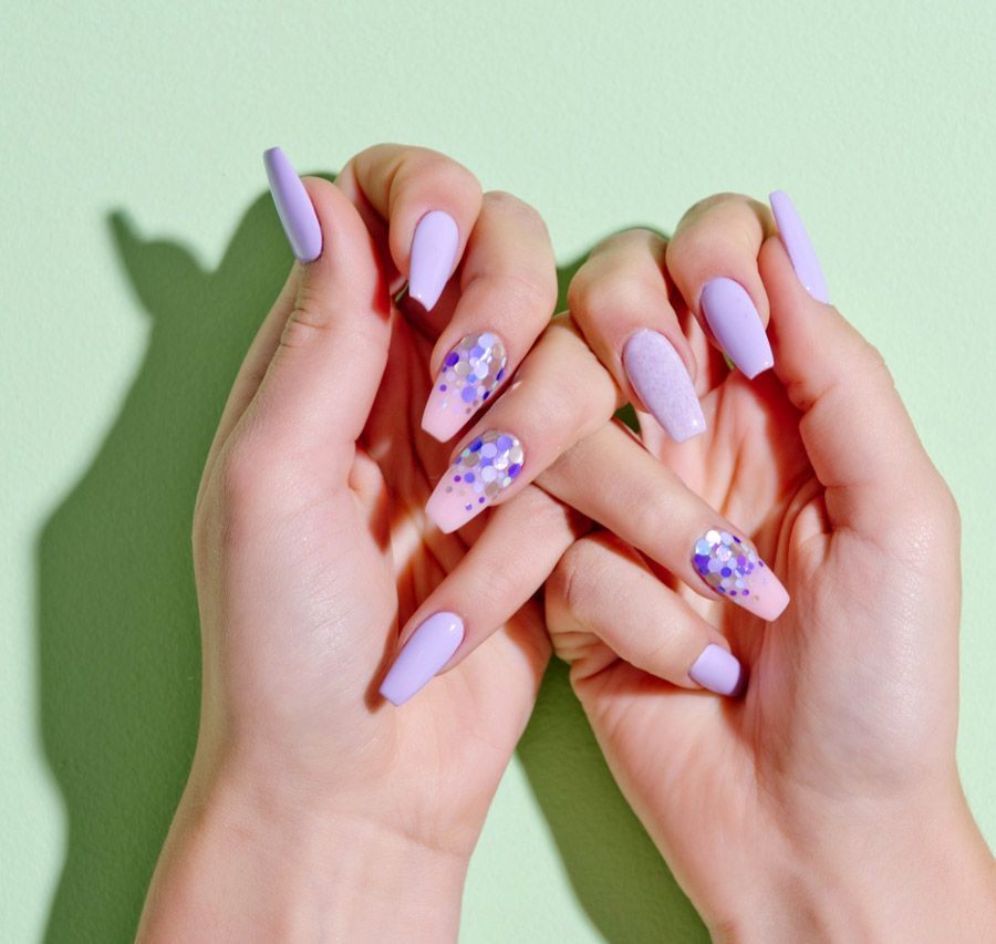 Manos con uñas en forma de ataúd pintadas de color lavanda y rosa degradado con detalles de purpurina morada sobre un fondo verde claro.