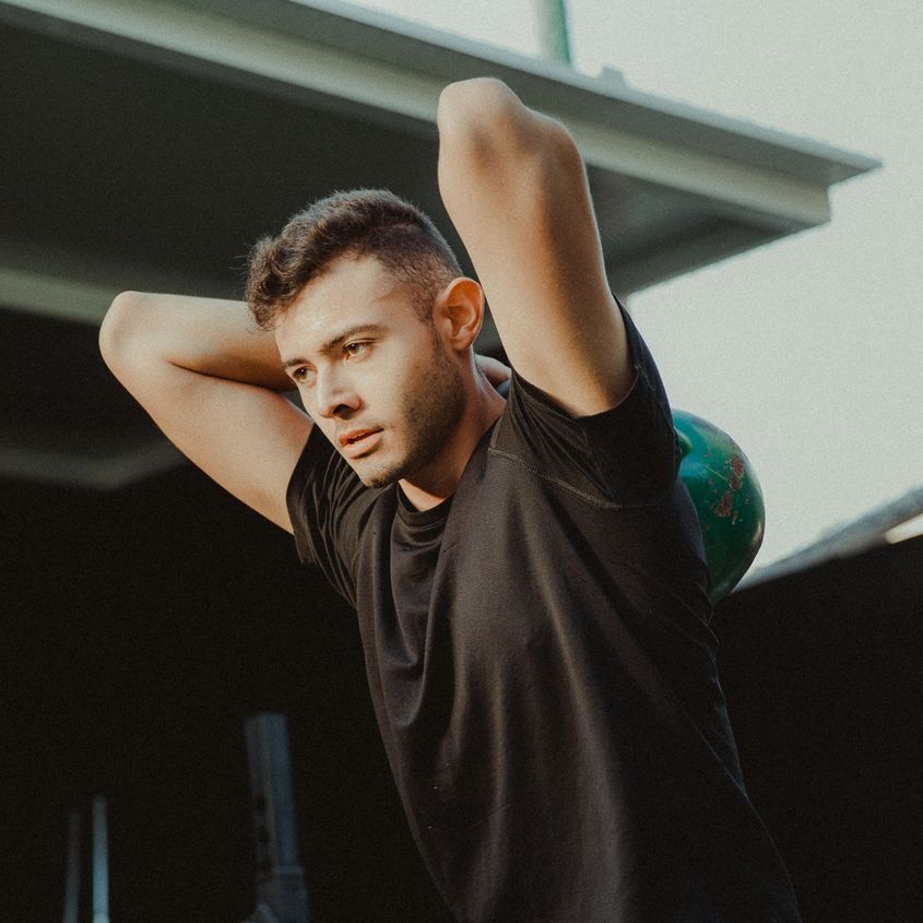 Un hombre con una camisa negra levanta una pesa rusa sobre su cabeza.