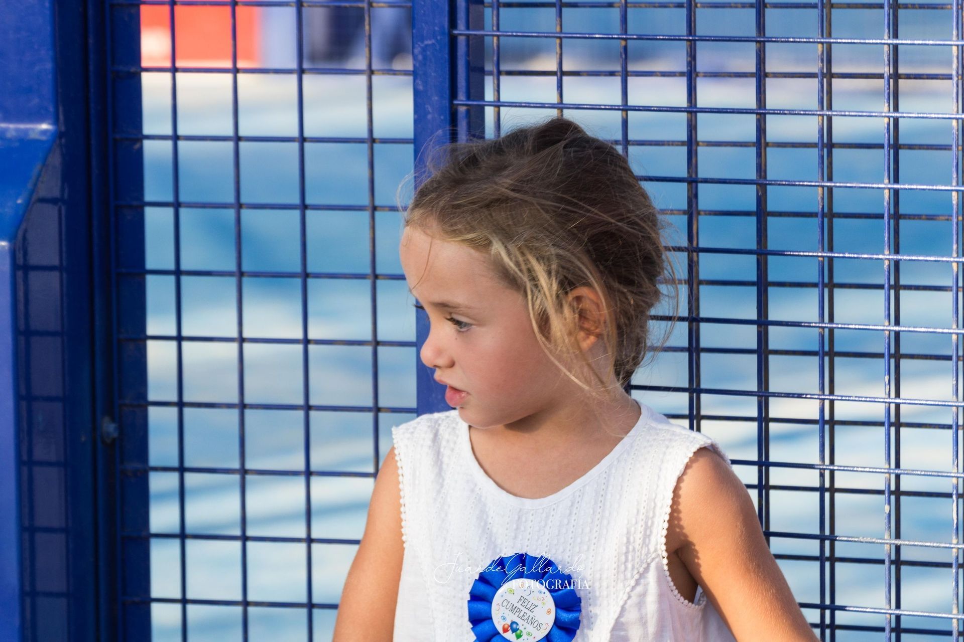 Una niña con un vestido blanco está sentada frente a una valla azul.